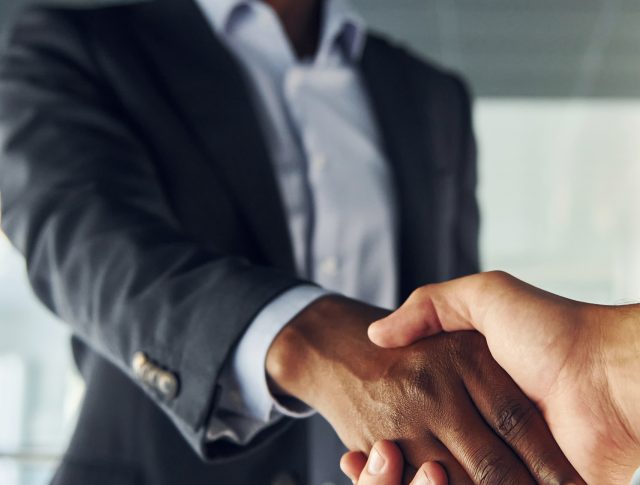 Doing handshake with customer. Young african american businessman in black suit is indoors.